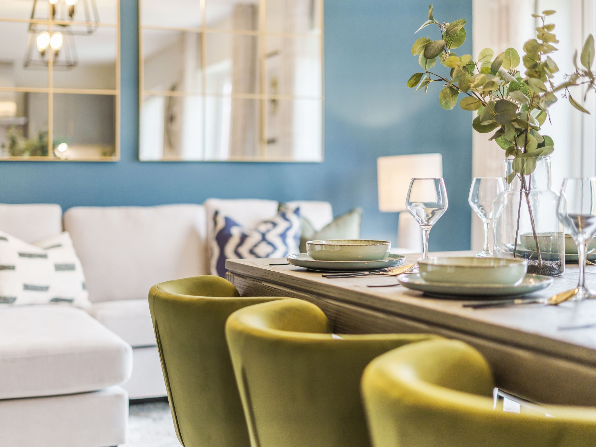 A bright and modern Charles Church dining room with a dining table and green chairs and a sofa in the background.