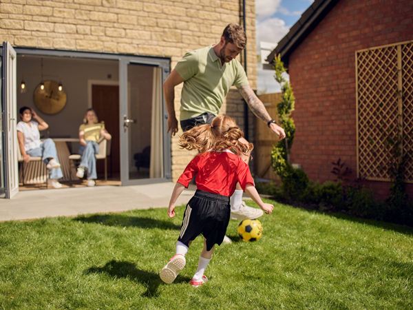 A father playing with a red headed child in a sports kit kicking a football on the grass of a modern new build Charles Church home.