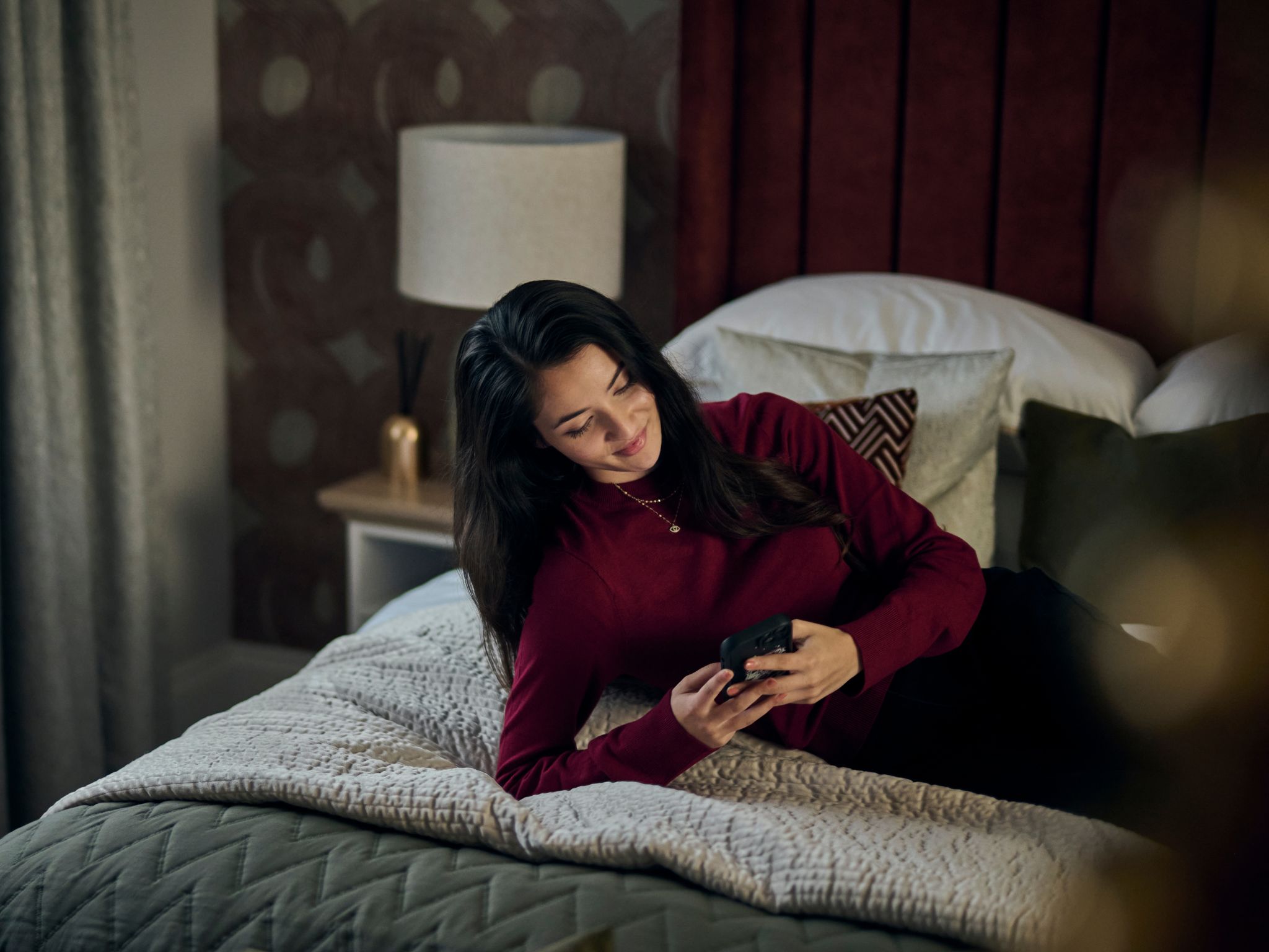 A woman reclining on a bed in a modern Charles Church home, smiling at a phone.