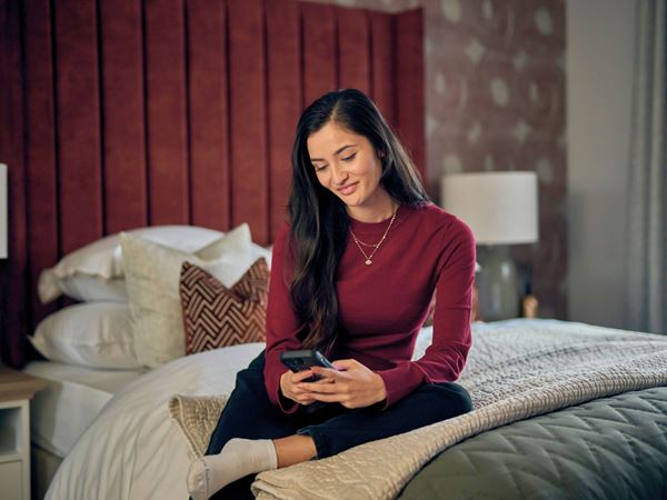 A woman wearing a red jumper sitting on a bed in a sultry Charles Church bedroom. 