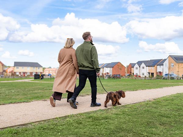 A man and a woman walking arm in arm along an open green space with their dog on the Charles Church Festival Park development.