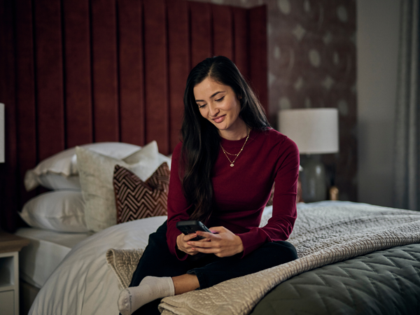 A woman sitting on a double bed with a burgundy headboard in a premium new build Charles Church home, looking at her phone.