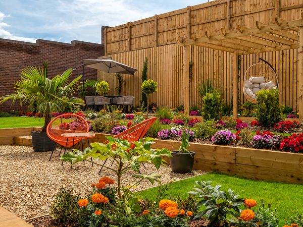 A beautiful garden with green grass and raised flower beds with pink and orange flowers, with two orange chairs and a small table on gravel.
