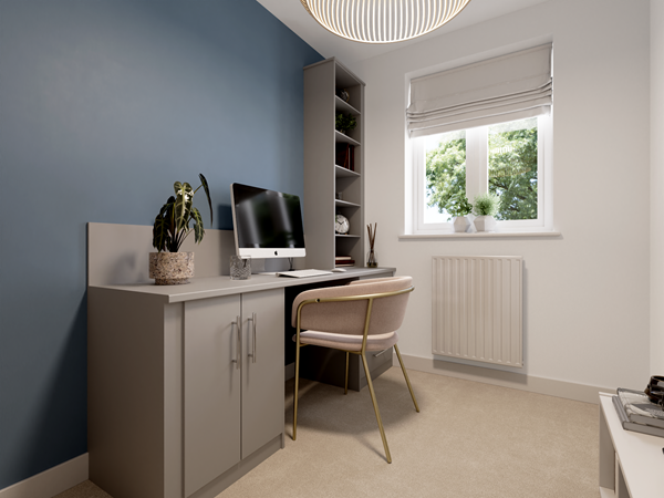 Compact home office with a gray desk, a computer monitor, and a pink upholstered chair with gold legs. A blue feature wall adds color, and a tall shelving unit holds books and decor. A window with a Roman blind provides natural light.