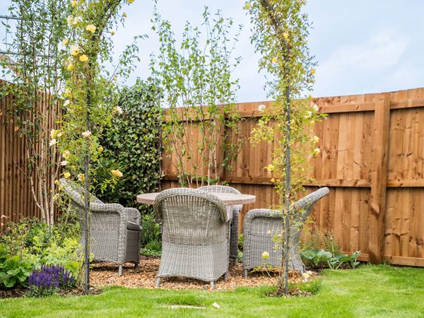 A round garden table with four rattan chairs in a garden with grass and beautiful flowers growing. 